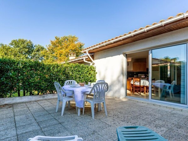 A patio with a table and chairs, a glass door leading to a kitchen, and a hedge in the background.