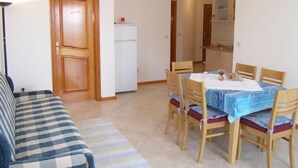 A living room with a sofa, a dining table with chairs, a refrigerator, and a kitchen area with cabinets and a sink.