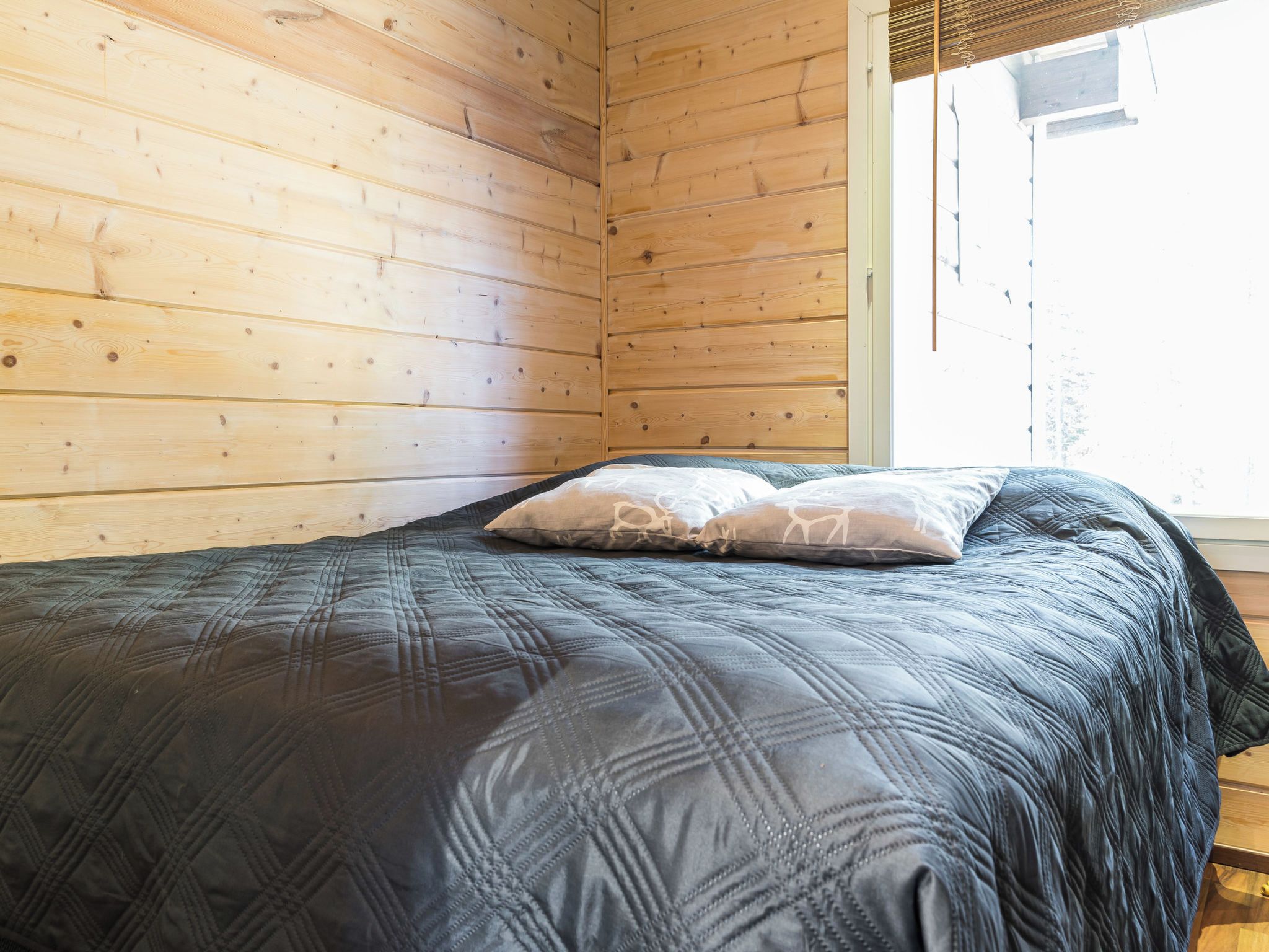 Un lit simple avec une couverture matelassée de couleur sombre et deux oreillers, dont l’un est orné d’un motif représentant un chien, dans une pièce aux murs en bois et dotée d’une fenêtre.