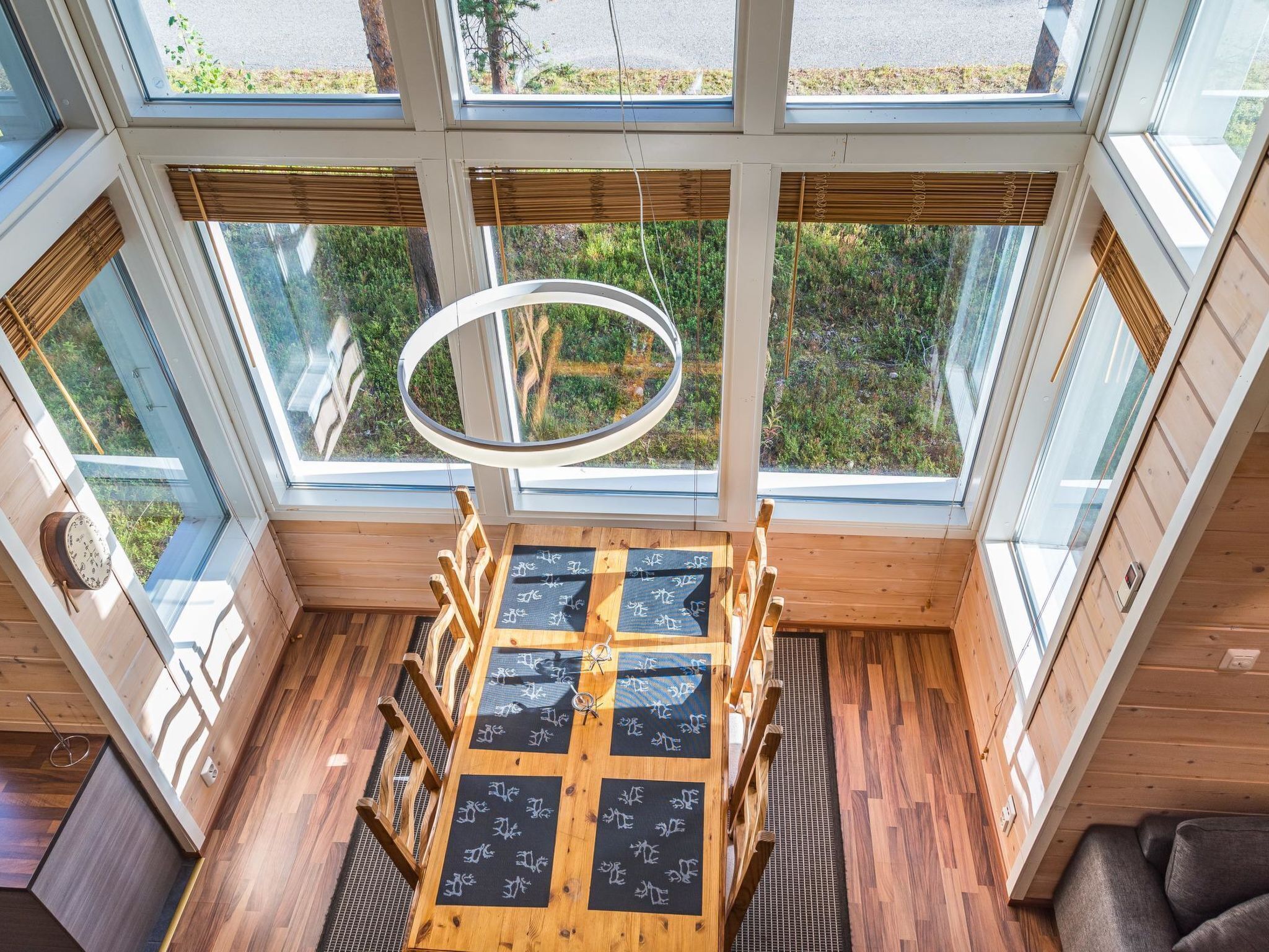 Quarto com amplas janelas, piso de madeira, luminária circular e um espaço de estar com mesa.