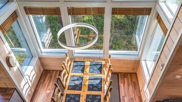 Quarto com amplas janelas, piso de madeira, luminária circular e um espaço de estar com mesa.