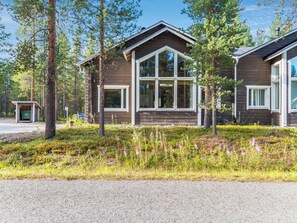 A cabin with a large window, surrounded by trees and a gravel driveway.
