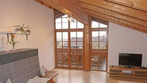 A spacious living room with a wooden ceiling, a large sliding glass door leading to an outdoor balcony, a flat-screen TV on a wooden stand, and a small table with a vase of flowers.
