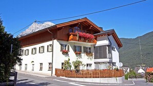 A two-story building with a balcony adorned with red flowers, a wooden facade, and a mountain backdrop.