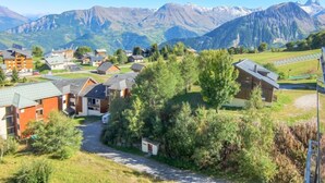 Un pueblo de montaña con casas de madera tradicionales, una calle central y montañas nevadas al fondo.