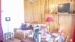 Una habitación acogedora con una pared de madera, un sofá con cojines estampados, una mesa de comedor con platos y una tetera, y un televisor sobre una estantería.