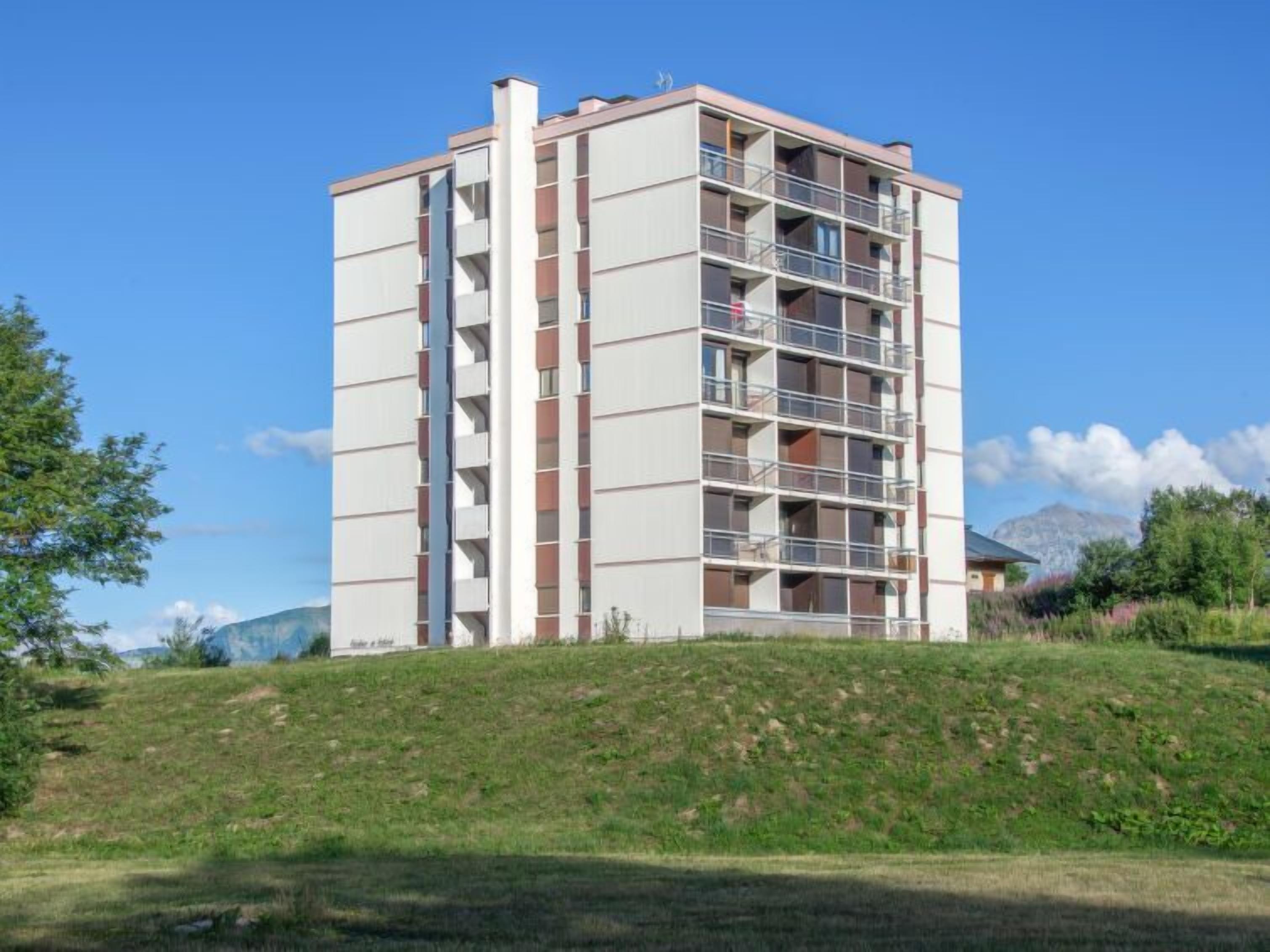 Un edificio residencial de varios pisos con balcones, ubicado en una colina cubierta de césped bajo un cielo despejado.