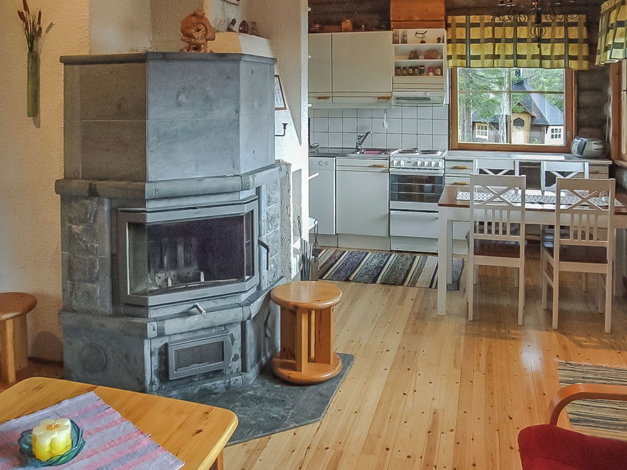 Ein Wohnzimmer mit Kamin aus Stein, Holzboden, einem Esstisch mit Stühlen, einer Küchenzeile mit weißen Schränken und einem Fenster mit Blick auf ein Haus.