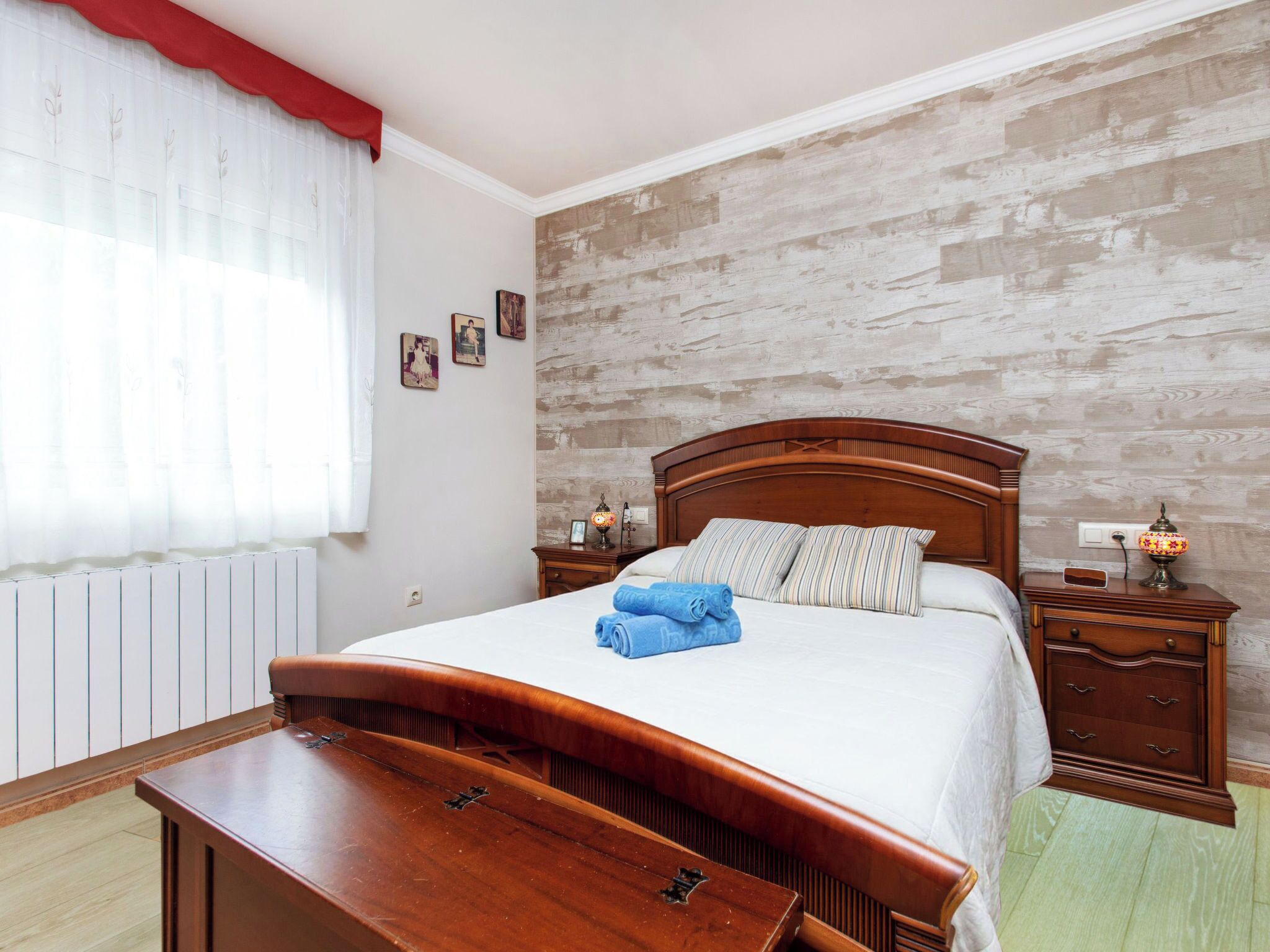 A bedroom with a wooden bed, a bedside table with a lamp, a radiator, and a window with sheer curtains.