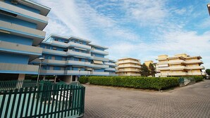 A multi-story residential building with balconies, a paved driveway, and a green fence. - Valbella by Interhome (Bibione)
