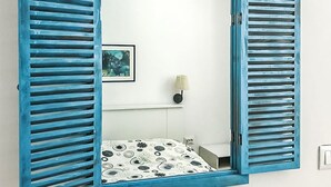 Een kamer met een grote spiegel, blauwe luiken, een witte muur, een ronde lamp en een bed met een decoratief beddegoed, plus een klein nachtkastje met een lamp.