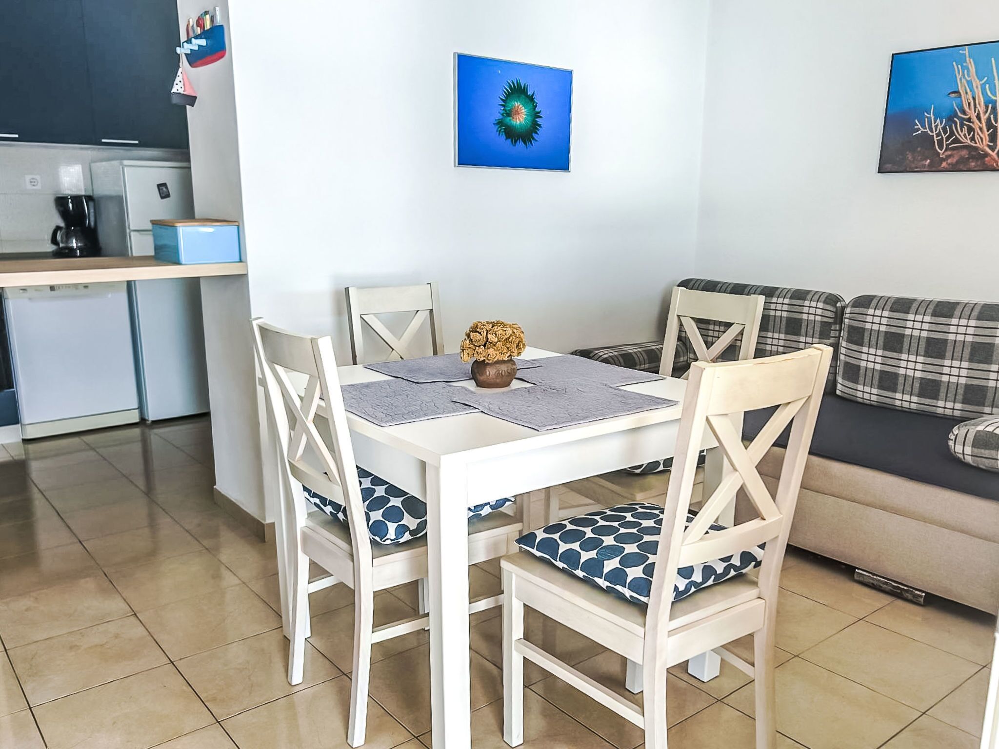 Un coin repas avec une table et des chaises blanches, une machine à café et un canapé agrémenté d’un coussin à carreaux.
