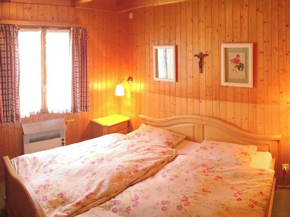 Una habitación de madera con una cama, una mesita de noche, una lámpara y un ventanal con cortinas.