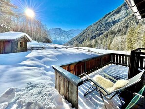 Una cabaña de madera con terraza y una silla, rodeada de nieve y montañas.