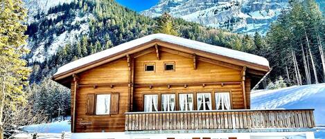 A wooden chalet with a balcony, set against a snowy mountain backdrop.