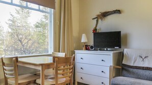 Quarto com uma mesa de jantar, cadeiras, cômoda branca com televisão, luminária e um galho decorativo na parede.