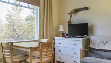 Een kamer met een eettafel, stoelen, een televisie op een dressoir, een lamp en uitzicht op bomen.