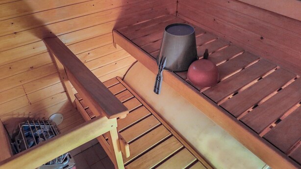 Uma sala de sauna com bancos de madeira, um banco com toalha, um balde de metal e uma bola vermelha.