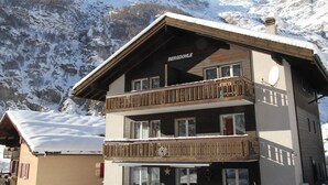 Un edificio de varios pisos con un balcón, con la leyenda “BERGDÖHLE”, frente a un paisaje montañoso cubierto de nieve.