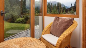 A wicker chair with a cushion and a view of a forest and mountains through glass doors. - Khamariah by Interhome (Villars)
