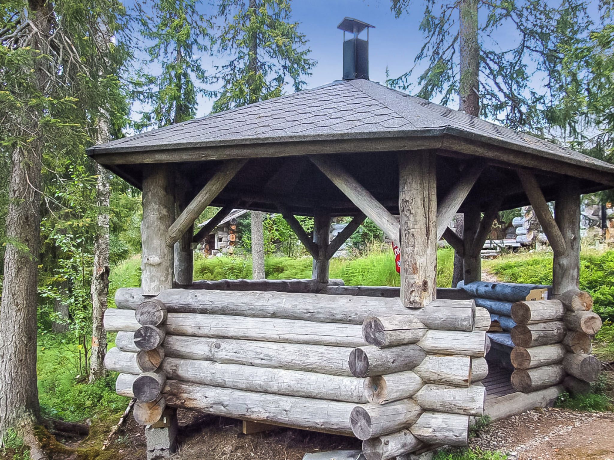 A wooden gazebo with a sloped roof, surrounded by logs and trees.