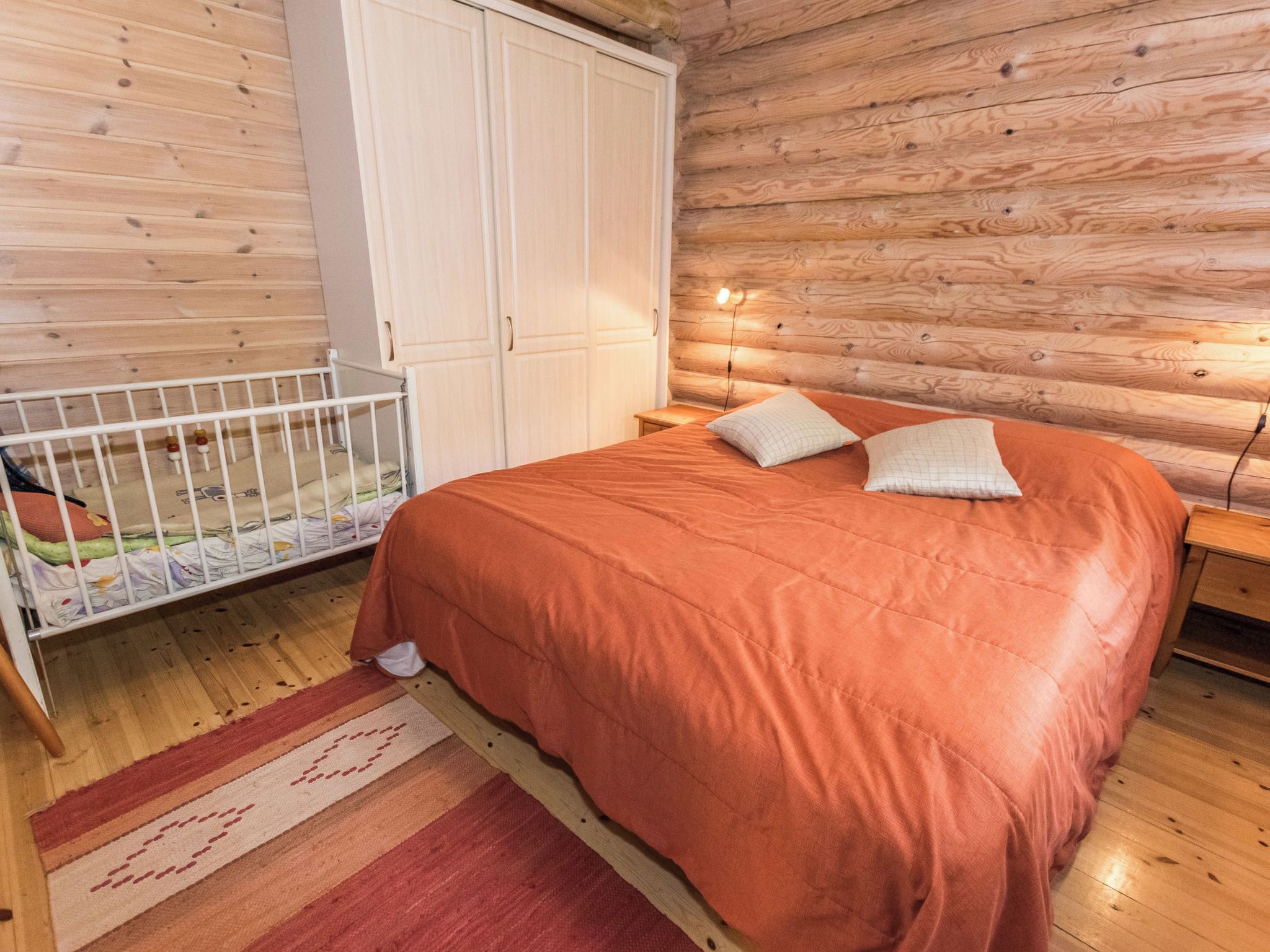A bedroom with a wooden floor, a bed with an orange bedspread, two pillows, a crib with a colorful mattress, and a white wardrobe.
