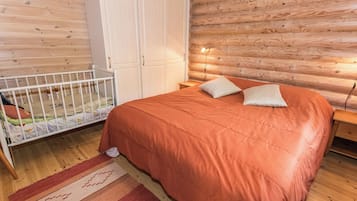 A bedroom with a wooden floor, a bed with an orange bedspread, two pillows, a crib with a colorful mattress, and a white wardrobe.