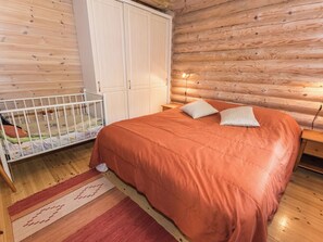 A bedroom with a wooden floor, a bed with an orange bedspread, two pillows, a crib with a colorful mattress, and a white wardrobe.