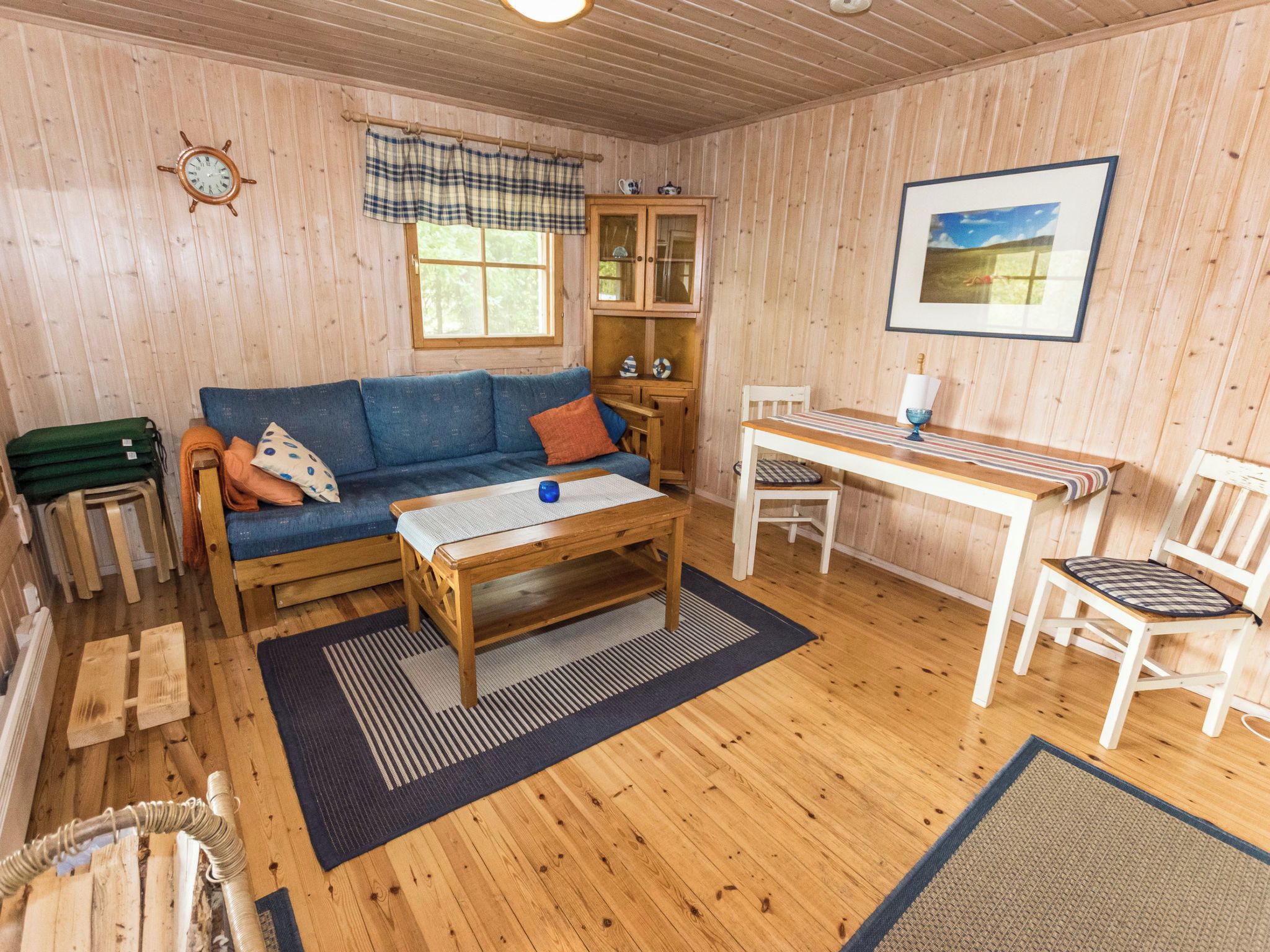 A cozy cabin interior with a wooden floor, a blue sofa, a wooden table, a white chair, and a window with a view of the outdoors.