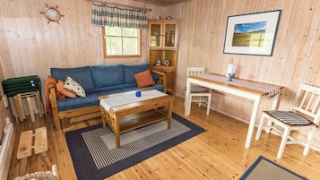 A cozy cabin interior with a wooden floor, a blue sofa, a wooden table, a white chair, and a window with a view of the outdoors.