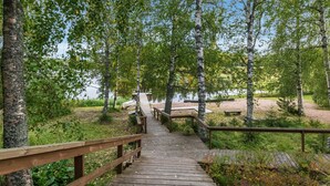A wooden boardwalk leading through a forested area with birch trees and a body of water in the background. - Koivuranta by Interhome (Jämijärvi)