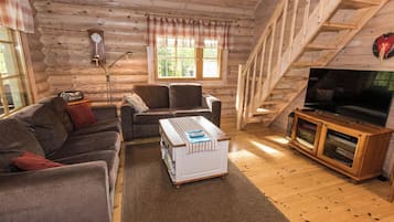 Interior acogedor de una cabaña con sofá, sillón, suelo de madera, televisor y un reloj en la pared.