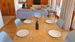 A dining area with a wooden table set for six, surrounded by light blue chairs. A bottle of wine and several wine glasses are on the table. In the background, there is a living room with a sofa and a television. - Hyacinthe 11 by Interhome (Villars)
