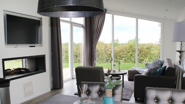 A modern living room with a flat-screen TV, a fireplace, a dining area with a tray, and a large window offering a view of greenery. - Green Resort Mooi Bemelen by Interhome (Bemelen)
