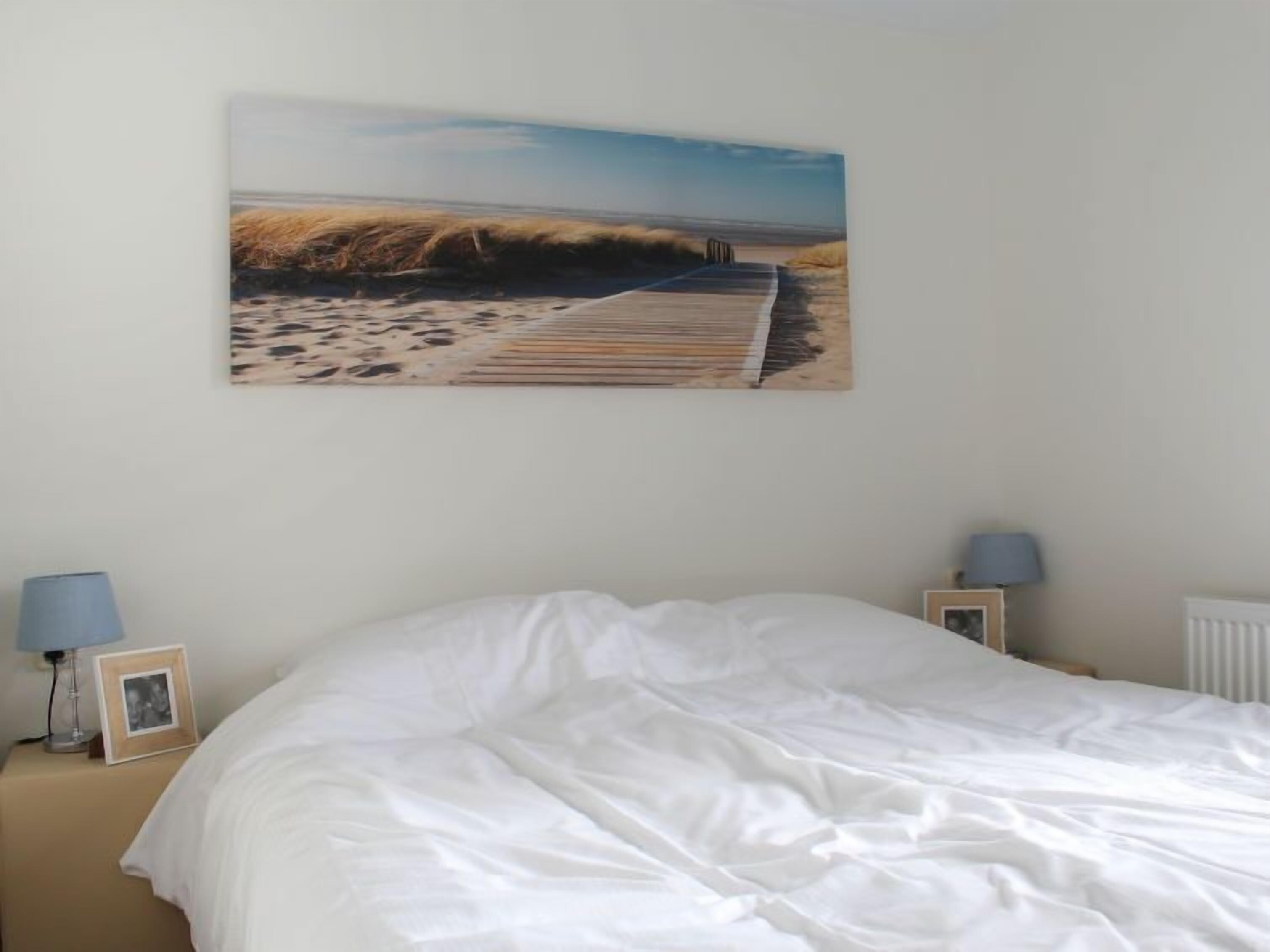 Ein Schlafzimmer mit einem Bett, einem Bild von einem Strandweg und zwei Nachttischlampen.