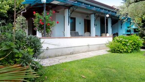Uma casa de dois andares com varanda coberta, jardim com diversas plantas e céu azul claro.