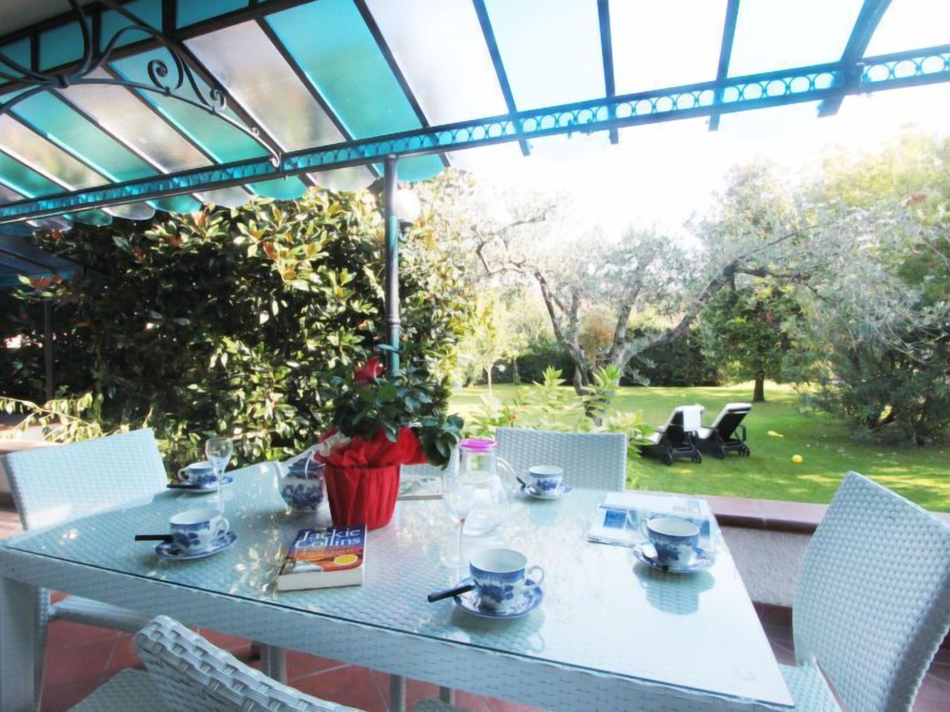 Une table à plateau en verre, sur laquelle sont posées des tasses à thé bleues et blanches, un livre et un vase rouge, le tout dans un décor de jardin.