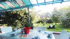 Uma mesa com tampo de vidro, xícaras de chá azuis e brancas, um livro e um vaso vermelho em um jardim.
