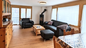Een gezellige woonkamer met een bank, fauteuil en voetenbankje, een houten tafel en een ingebouwde houtkachel.