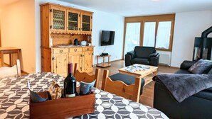 Een gezellige woonkamer met een houten kast, een zwarte fauteuil, een bank, een eettafel met een tafelkleed met patroon, en een fles wijn op de tafel.