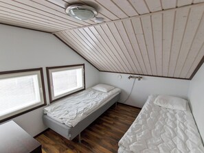 A small, single-bed room with wooden flooring, a sloped ceiling, and two windows with blinds.
