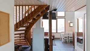 A spacious living area with a wooden staircase, a dining area with white chairs, and large windows allowing natural light.