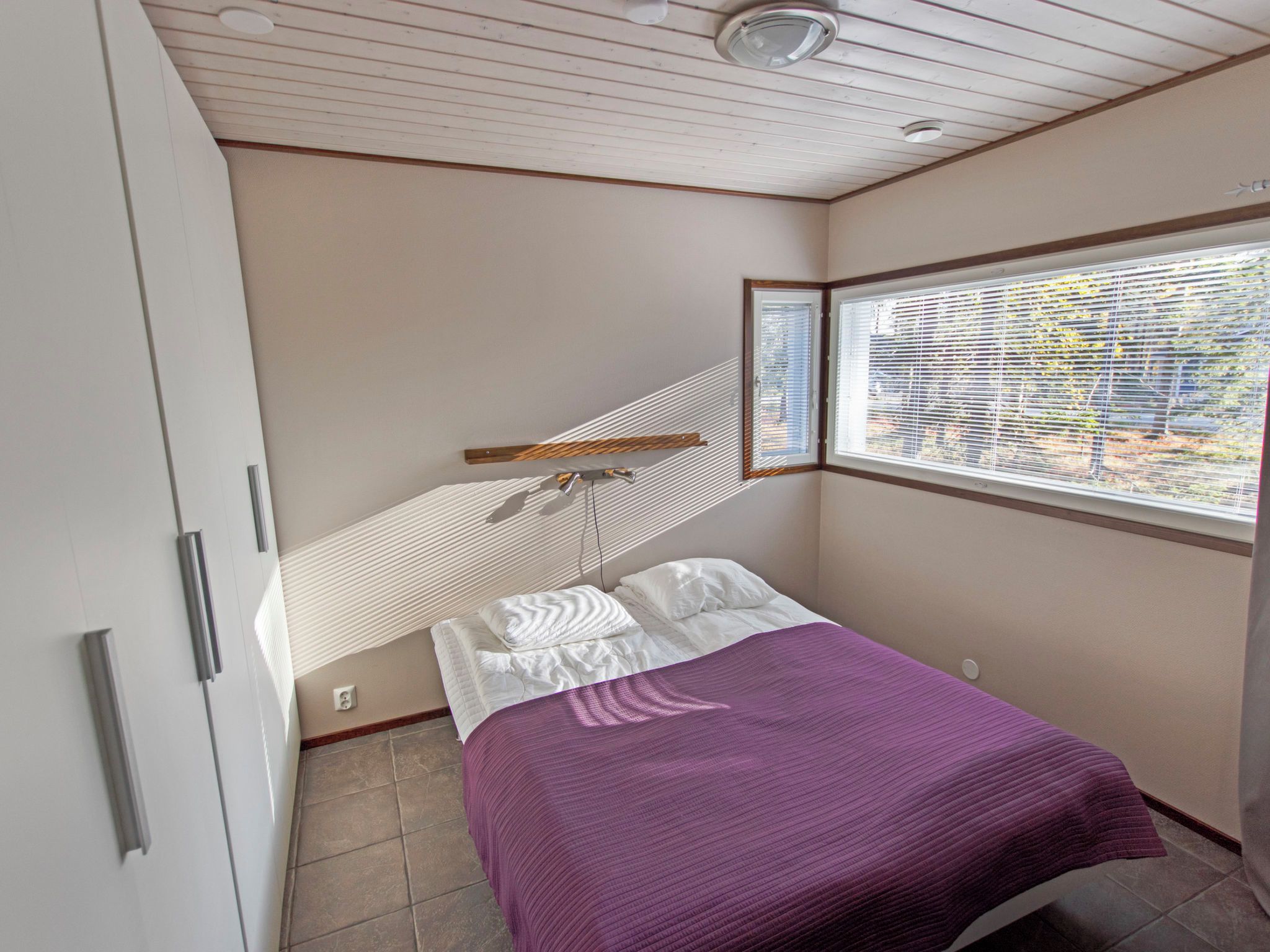 Une chambre à coucher avec un lit, une fenêtre avec des stores, une tête de lit en bois et un ventilateur de plafond.