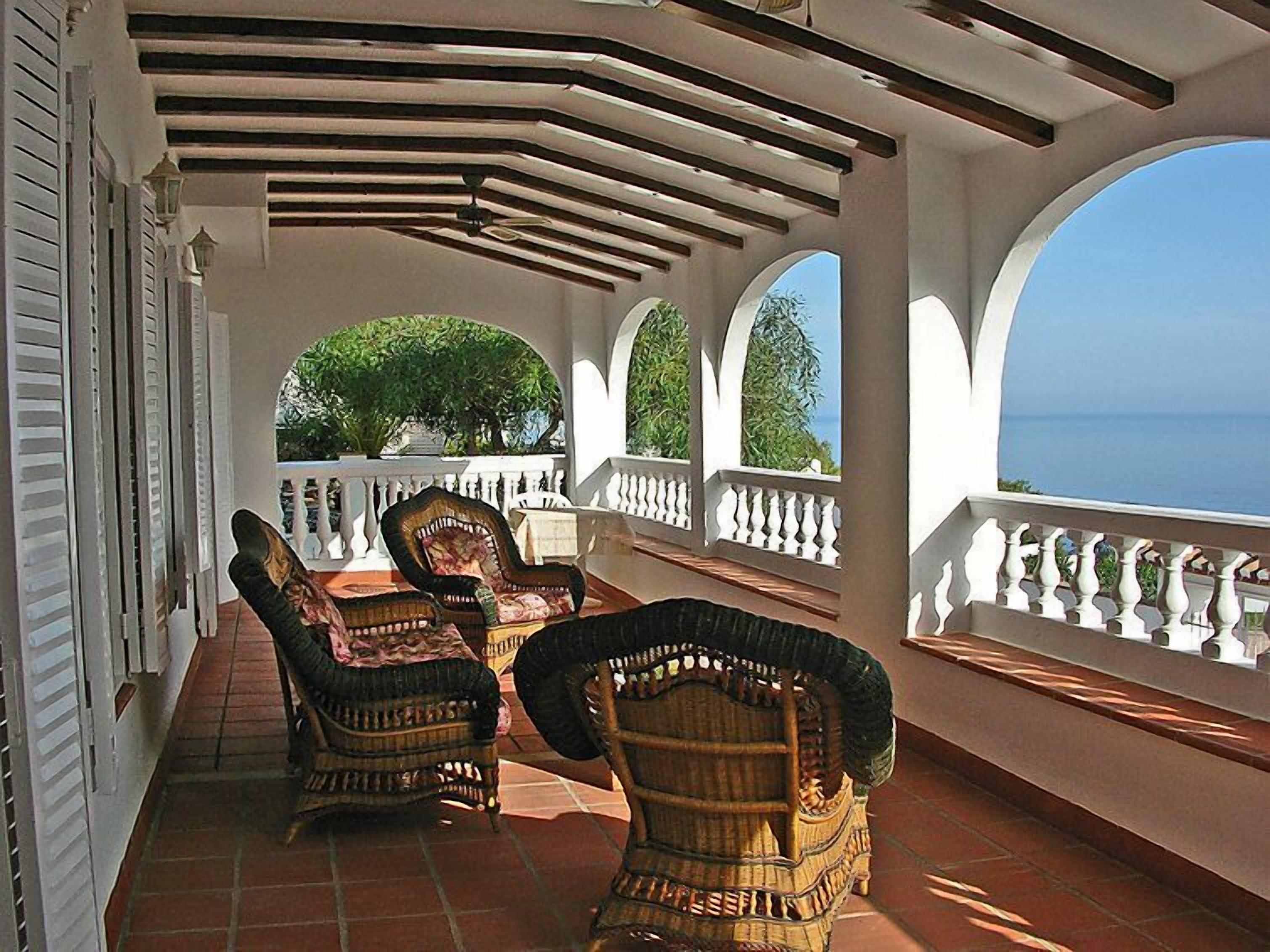 A covered outdoor seating area with wicker chairs and a view of the sea.