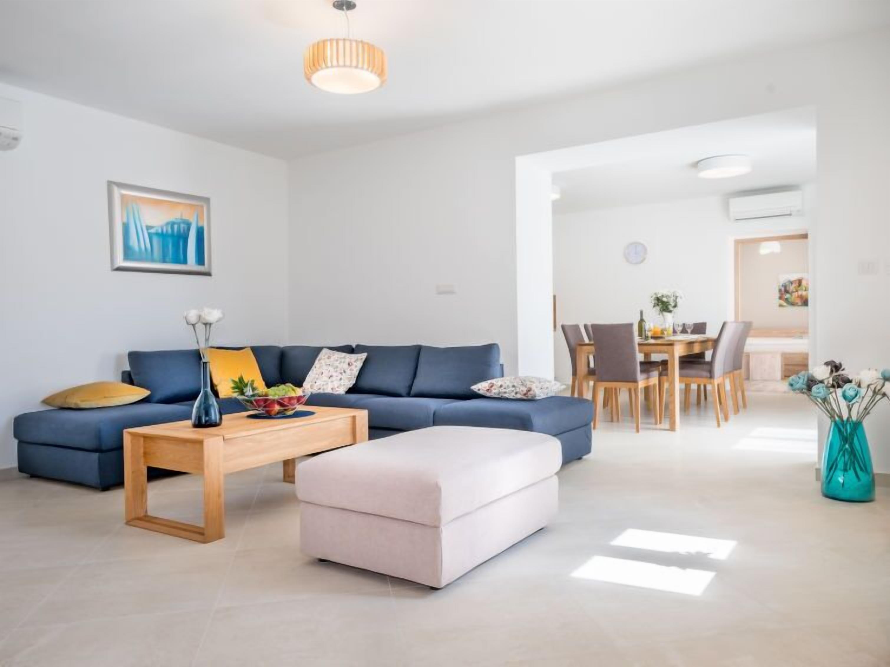 A modern living room with a blue sofa, a wooden coffee table, and a white ottoman. There is a dining area with a wooden table and chairs.