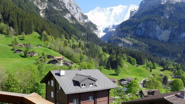 Ein Bergdorf mit traditionellen Holzhäusern, freier Aussicht auf die umliegenden Berge und einer kleinen Holzdielen-Terrasse im Vordergrund.