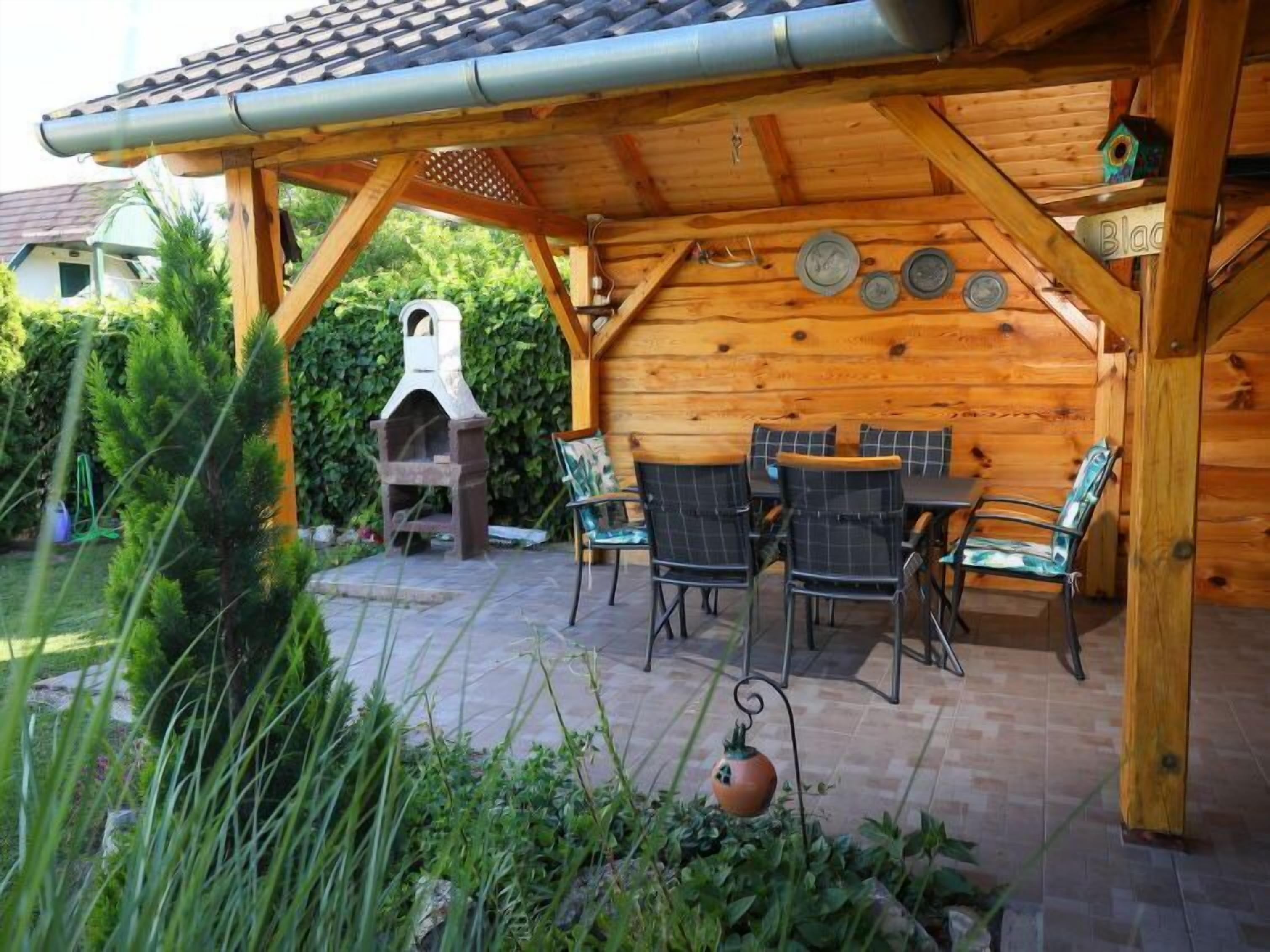 A wooden outdoor patio with a stone barbecue grill, a wooden table with chairs, and a decorative birdhouse.