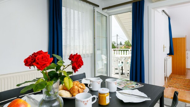 Spiseområde med et bord dækket til morgenmad, inklusive croissanter, æbler og en vase med røde roser.