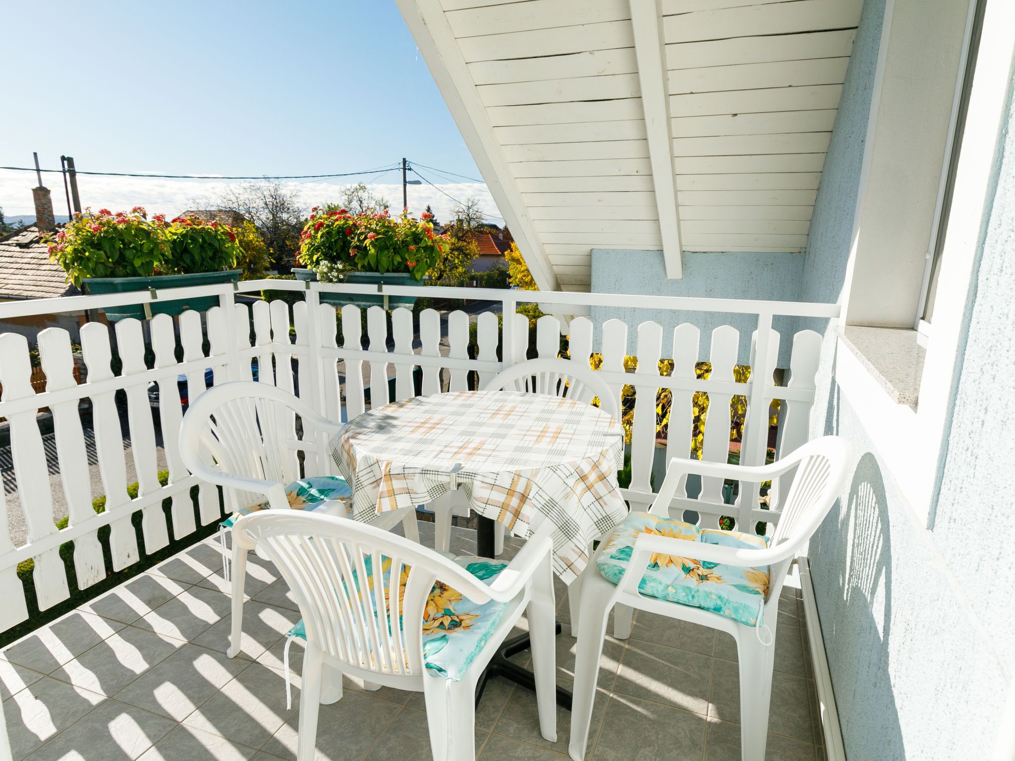 A balcony with a white picket fence, a round table with a plaid tablecloth, and white plastic chairs with colorful cushions.