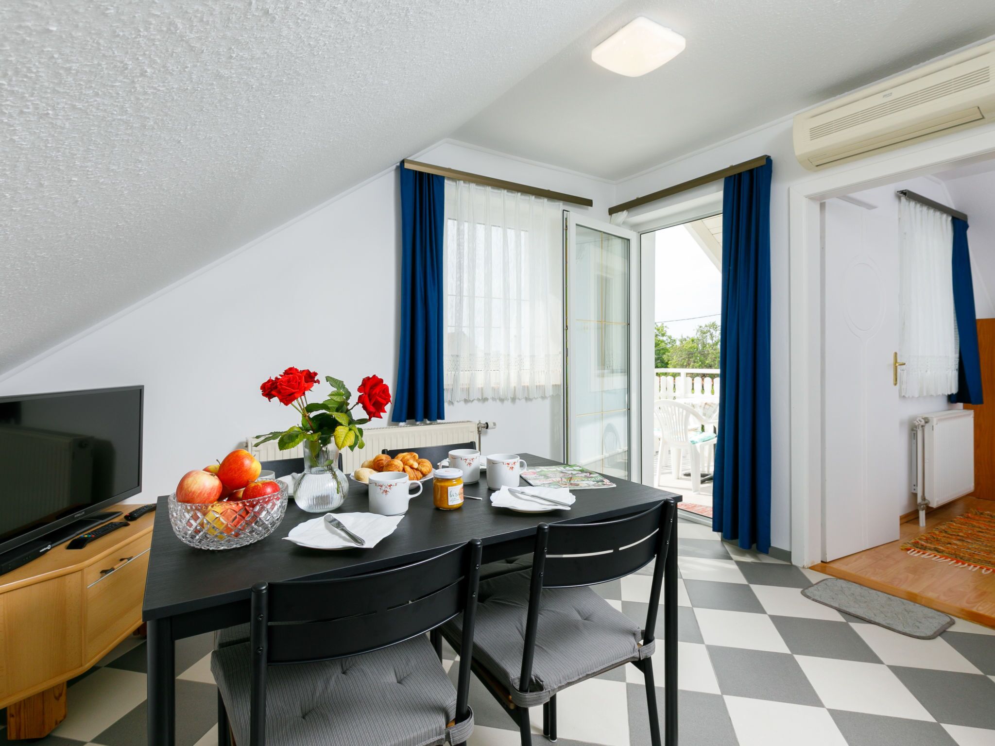 A dining area with a table set for two, a TV, and a vase with flowers.